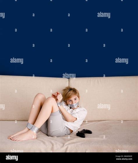 Blonde Boy On The Sofa Wrapped In Self Adhesive Duct Tape And With His Mouth Taped Next To The