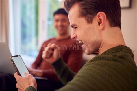 Same Sex Male Couple On Sofa At Home Using Mobile Phone And Laptop