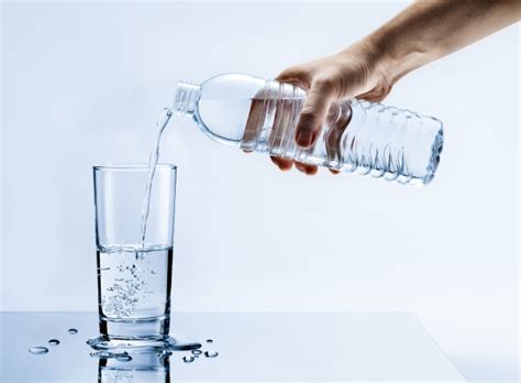 Рука наливает свежую чистую воду из бутылки в стакан на столе с каплями ...