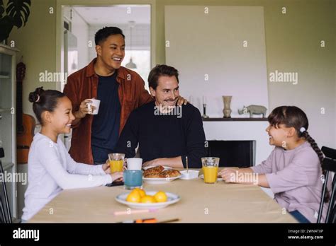 Happy Gay Couple Having Fun While Having Breakfast With Daughters At Dining Table In Home Stock