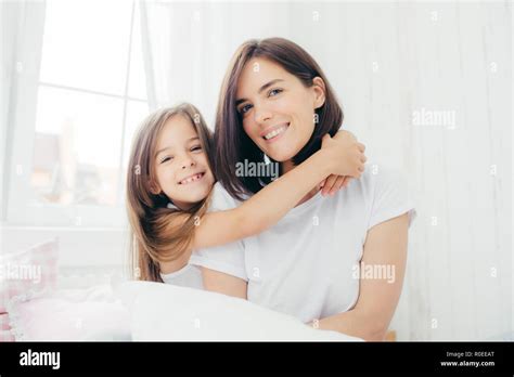 Indoor Shot Of Good Looking Brunette Mother With Gentle Smile And Her Small Daughter Gives Hug