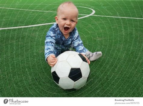 Little boy sitting on green grass of playing field with soccer football ball and looking at ...