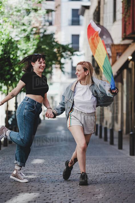 Cheerful Lesbians Holding Hands And Waving Lgbt Flag While Walking On Blurred Background Of City