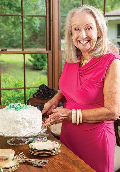 An Older Woman In A Pink Dress Cutting Into A Cake On Top Of A Wooden Table