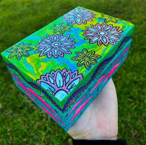 A Hand Holding Up A Painted Box In The Grass With Flowers On Its Sides