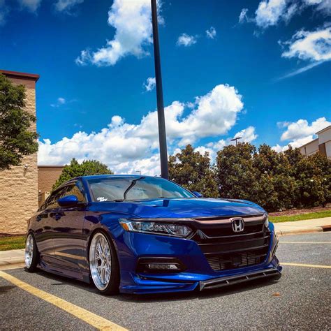 A Blue Car Parked In A Parking Lot Next To A Tall Pole With Clouds Above It