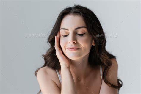 Smiling Brunette Woman Closing Eyes And Touching Face Isolated On Grey