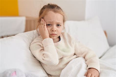 Adorable Blonde Girl Touching Head For Headache Sitting On Bed At