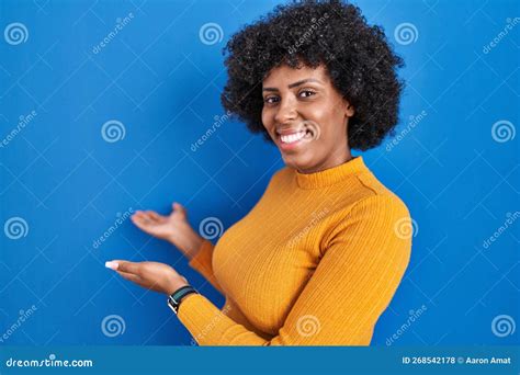 Black Woman With Curly Hair Standing Over Blue Background Inviting To Enter Smiling Natural With