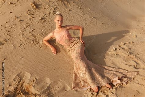 Blonde Woman In Splendid Gold Dress Posing In Desert Lying On The Sand At The Sunset Stock