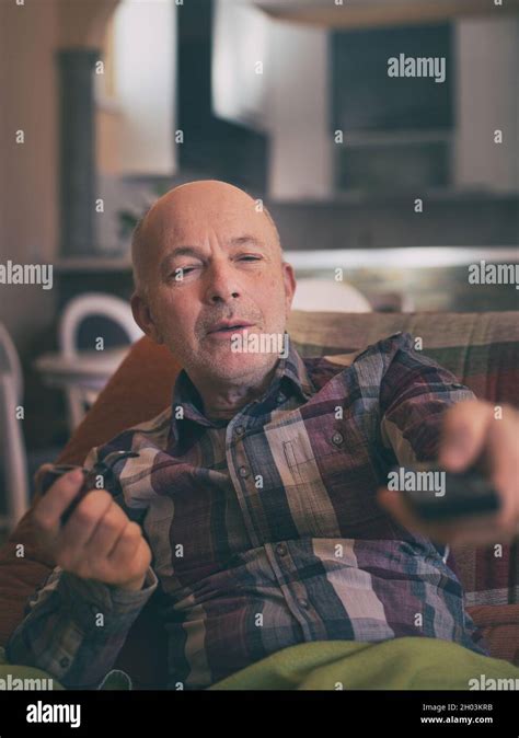 Mature Man Sitting On Sofa At Home Holding Remote Control Smoking Pipe