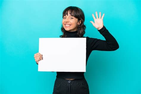 Joven latina aislada de fondo azul sosteniendo un cartel vacío y saludando Foto Premium