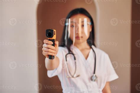 Holds infrared thermometer. Young serious asian woman standing indoors