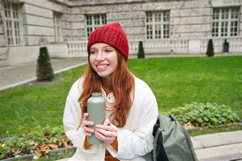 Smiling Redhead Girl Rests In Park Sits On Bench With Backpack Drinks From Thermos Enjoys Hot