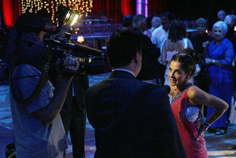 A Woman In A Red Dress Standing Next To A Man With A Camera On His Shoulder