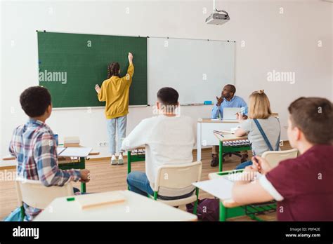 Rear view of schoolgirl writing on chalk board and classmates with teacher sitting in classroom ...