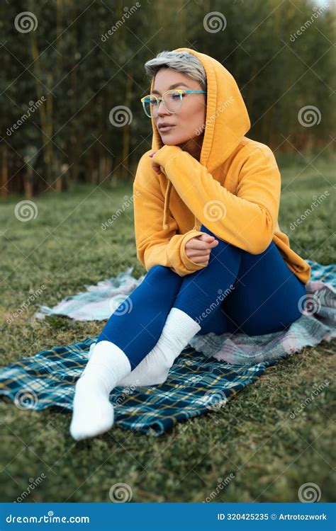 Cute Girl With Glasses In An Orange Hoodie Is Sitting In A Park In