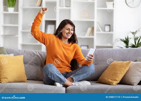 Excited Young Indian Woman Sitting On Couch Holding Smartphone Stock